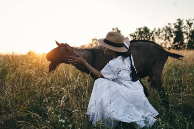 Tarladaki genç kadın keçilerle yürüyor. Kız ve keçiler yazın çayırda. Hayvanlar için sevgi. Keçi çiftliği. Evcil hayvanlar. Hayvanlı mutlu kadın. Hayvanlara şefkat ve sevgi. Evcil bir hayvanı öpüyor.