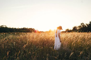 Yaz tatili ve seyahat. Kırsal keten kumaş içinde kaygısız genç bir kadın yaz çayırlarında dinleniyor. Gün batımında kır çiçeklerinin arasında poz veren hasır şapkalı şık bohem kadın. Atmosferik an