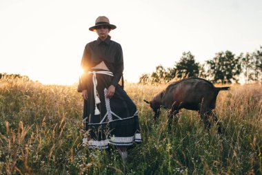 Genç kadın tarlada beyaz keçilerini otlatıyor. Kız ve keçiler yazın çayırda. Hayvanlar için sevgi. Keçi çiftliği. Evcil hayvanlar. Hayvanlı mutlu kadın. Hayvanlara şefkat ve sevgi. Evcil bir hayvanı öpüyor.
