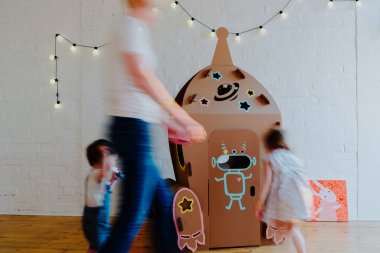 Neşeli kardeşler birbirlerinin arkasından koşuyorlar ve karton roket etrafında oturma odasının ortasında oyun oynarken duruyorlar.