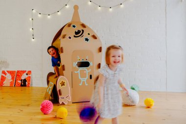 Neşeli kardeşler birbirlerinin arkasından koşuyorlar ve karton roket etrafında oturma odasının ortasında oyun oynarken duruyorlar.