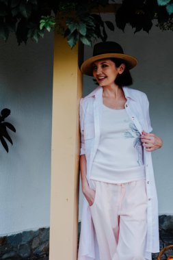 Gündelik, açık renkli elbiseler ve geniş kenarlı bir şapka giymiş bir kadın yeşil bir gölgeliğin altında direğe yaslanıyor..