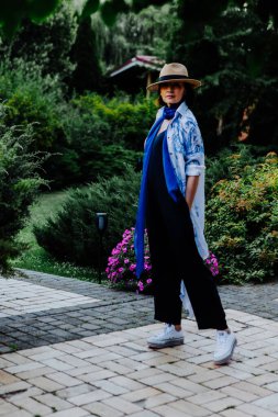 Havalı bir kadın, uzun, dalgalı desenli bir ceket, mavi bir eşarp, geniş bacaklı siyah pantolon, beyaz spor ayakkabılar ve hasır bir şapkayla dışarıda dolaşırken görülüyor.