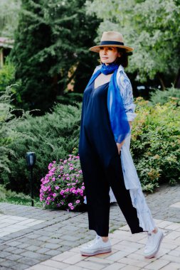 Havalı bir kadın, uzun, dalgalı desenli bir ceket, mavi bir eşarp, geniş bacaklı siyah pantolon, beyaz spor ayakkabılar ve hasır bir şapkayla dışarıda dolaşırken görülüyor.