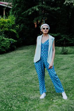 A fashionable woman stands confidently on a green lawn, wearing a bold, geometric-patterned blue jumpsuit, a light sleeveless vest, and white sneakers. clipart