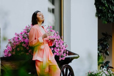 Serene kadın açık havada canlı bir çiçek çalısının yanında duruyor, ipeksi pembe bir bornoz giymiş ve elinde çay fincanı tutuyor.