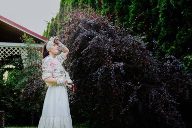 Woman dressed in a floral gardening outfit is pruning a large shrub in a lush garden. clipart