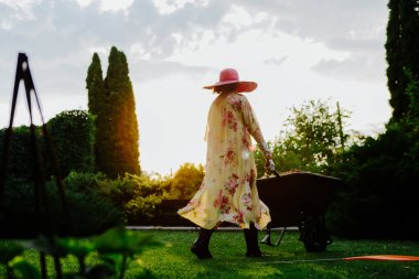 Çiçekli bir yaz elbisesi ve geniş kenarlı güneş şapkası giyen bir kadın gün batımında bir bahçeyle ilgilenirken görülüyor. Uzun ağaçlar ve yemyeşil ağaçlarla çevrili güzel manikürlü bir çimenlikte el arabasıyla yürüyor.