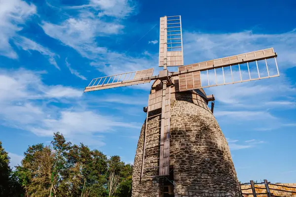 stock image Old stone windmill stands against a clear blue sky with gentle clouds. The windmill wooden blades and stone structure showcase traditional craftsmanship and the historical significance of rural architecture.