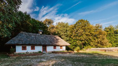 Geleneksel sazdan çatılı ev, berrak mavi gökyüzünün altında yemyeşil bir manzarayla çevrili kırsal bir ortamda huzur içinde oturur. Beyazlatılmış duvarlar ve kırsal ahşap pencereler kırsal yaşamın basitliğini ve güzelliğini vurgular..