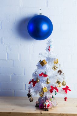 Small white Christmas tree decorated with golden and red ornaments, pine cones, and miniature Santa figurines on a wooden table against a white brick wall. clipart