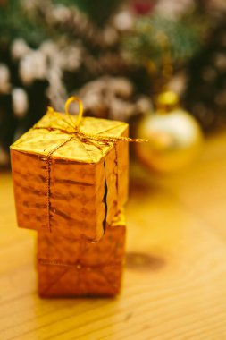 Tahta bir yüzeye istiflenmiş küçük altın hediye kutuları, şenlikli bir Noel çelenginin bulanık arka planına karşı..