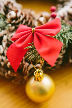 Christmas Decoration. Christmas wreath made of pine cones, decorated with red bows, evergreen branches, and gold ornaments, sits on a wooden surface. clipart