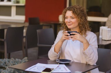 Kıvırcık saçlı bir kadın açık bir kafede otururken bir fincan kahveden hoşlanır. Etrafı dokümanlar ve akıllı telefonlarla çevrili. Bu da modern, kentsel bir ortamda iş ve rahatlama karışımını gösteriyor..