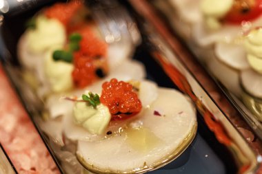 Elegant seafood appetizer featuring thinly sliced scallops, garnished with vibrant salmon roe, cherry tomatoes, microgreens, and a delicate sauce, artistically presented on sleek plates.