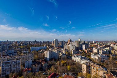Geniş hava manzarası Kyiv 'in dinamik ufuk çizgisini açık mavi bir gökyüzü altında yakalıyor, modern yüksek binalar ve işlek bir şehir caddesinin yanında tarihi binaların canlı bir karışımını sunuyor.