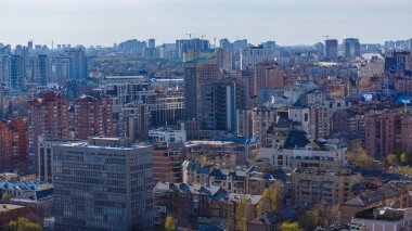 Bulanık bir gökyüzü altındaki Kyiv kentinin geniş hava manzarası, inşaat vinçleriyle noktalanmış modern ve eski binaların yoğun bir karışımını içeriyor.