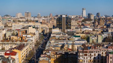 Geniş açılı canlı manzara Kyiv 'de işlek bir caddeyi yakalıyor, açık mavi gökyüzü altında aktif trafiği olan tarihi ve modern binaların dinamik bir karışımını gösteriyor.