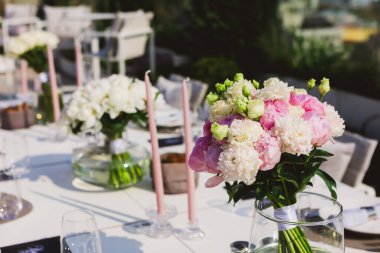 Zarif bir açık masa seti pembe ve beyaz şakayık buketleri ile süslenmiş, narin mumlar ve cam süslemeler ile tamamlanmış, kutlama, romantizm, lüks etkinlikler ve sofistike toplantılar temaları çağrıştıran.