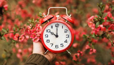 Çiçeklerin içinde retro kırmızı alarmlı bir saati tutan el. İlkbahar, ilkbahar, yaz saati tasarruf zamanı afişi veya arkaplan. 