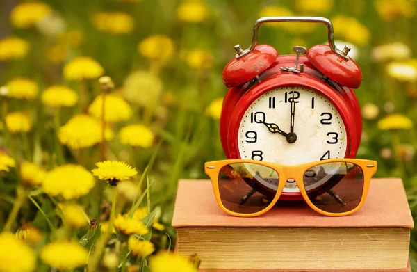 草の中に黄色い花の本に目覚まし時計とサングラス 学校の終わり夏休みの時間 — ストック写真
