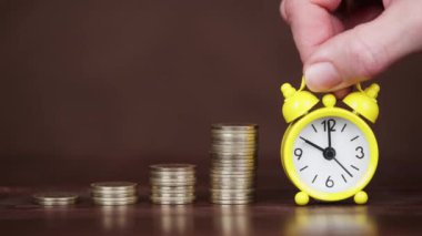 Bir el, altın para yığınlarının yanında çalar saati tutar. Zaman paradır, tasarruf geçmişi.