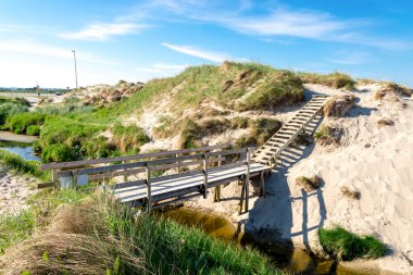 A wooden boardwalk bridge over a narrow stream to a small sand dune near Sola beach, Stavanger, Norway, May 2018