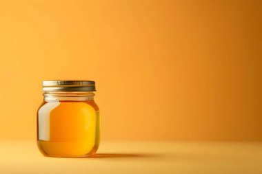 Altın renkli bir sıvıyla dolu şeffaf cam bir kavanoza yakın plan, muhtemelen bal ya da şurup, parlak, minimalist bir stüdyo arka planına ayarlanmış. Kavanozun içindekiler parıldıyor ve parlıyor..