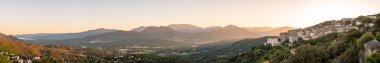 Panoramic view of early morning sunlight flooding the valley and distant Bavella mountains behind the town of Sartene in Corsica with bay of Propriano in the distance clipart