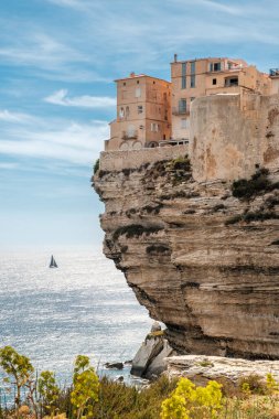 Korsika 'daki Bonifacio Kalesi' nde deniz ve hava koşulları altında kireçtaşı kayalıklara tünemiş evler. Uzakta yelkenli bir yat var.