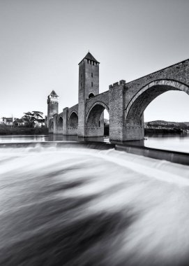 Fransa 'nın Cahors kentindeki River Lot' u geçen bir UNESCO Dünya Mirası Alanı olan 14. ve 15. yüzyıl Valentre köprüsü altında akan suyun siyah ve beyaz görüntüsü.