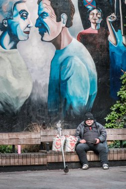 Londra, İngiltere - 28 Nisan 2025: Parktaki bankta güvercinli evsiz bir adam, Londra 'daki Tottenham Court Road açıklarındaki Whitfield Garden' da büyük boyalı bir duvar resminin altında telefonunu kontrol ediyor.