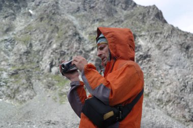 Göze çarpan turuncu ceketli cesur bir adam, ustalıkla dağlık manzaranın nefes kesici görüntülerini yakalıyor. Kamerasını kullanarak zorlu bir kayanın üzerinde cesurca duruyor..