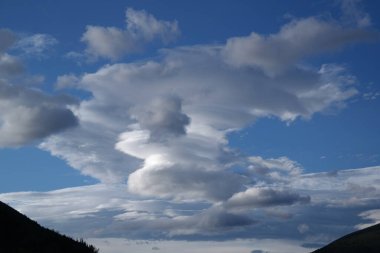 İnce beyaz bulutlar görkemli karanlık dağların üzerinde süzülür. Derin mavi gökyüzünün altında yemyeşil ağaçlarla çevrili. Güneş ışıl ışıl parlıyor, huzurlu ve huzurlu bir atmosfer yaratıyor..