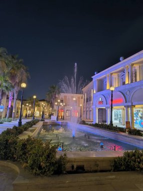 Geceleri şehir meydanında merkezi bir çeşme, çevredeki dükkanlar ve restoranlar parlayan ışıklarla süslenir. Palmiye ağaçları, karanlık ve berrak bir gökyüzünün altındaki geçitleri kaplıyor..