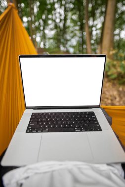 Design mock-up of a modern laptop computer with a blank white screen. Hiker is resting in a hammock in the woods. Using computer outdoors concept.