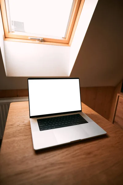Opened modern professional laptop on a wooden table against opened roof window. Concept of working remotely in a cottage. Design mock-up with blank computer screen.
