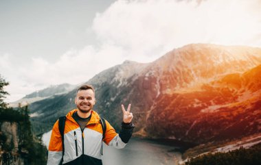 Dağın tepesinde sakallı, beyaz bir adam var. Arkaplan dağ manzarası ve canlı günbatımında göl. Gölün üzerindeki kayalıklarda duran bir adam. Başarı, özgürlük ve güç kavramı.