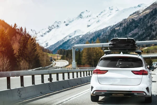 Black Roof Box on a Sporty White SUV Family Car. Rooftop cargo carrier bag. Rear view of a car with a roof box. Alpine highway. Removable black car trunk for luggage on the roof of a car.