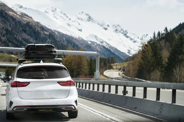 Rooftop cargo carrier bag. Rear view of a car with a roof box. Black luggage compartment box on a sport white car, back view. Alpine highway. Black Roof Box on a Sporty White Wagon Family Car