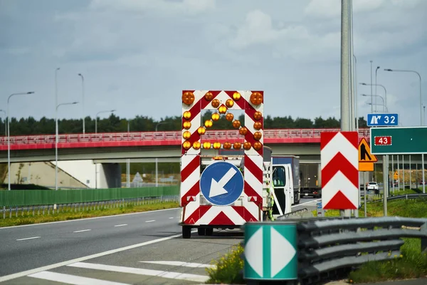 Trafiği sol şeride yönlendiriyor. Karavanda sarı ok işareti var. Bir yol işyeri aracı ve bir işçi otoyoldaki inşaat malzemeleriyle meşguller..