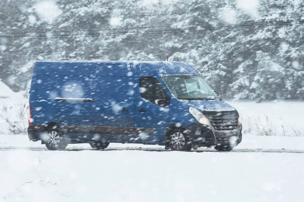 A van transporting cargo on a snowy road in winter.Side view of a blue van driving on a road covered with snow and ice. The road is surrounded by a snowy landscape and a cloudy sky.