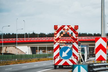 İşaret levhası parlayan bir okla trafiği otoyoldaki farklı şeritlere yönlendirir. Bir servis aracı ve bir işçi tabelasının arkasında yol tamiri üzerinde çalışıyor. Yol çalışmaları.