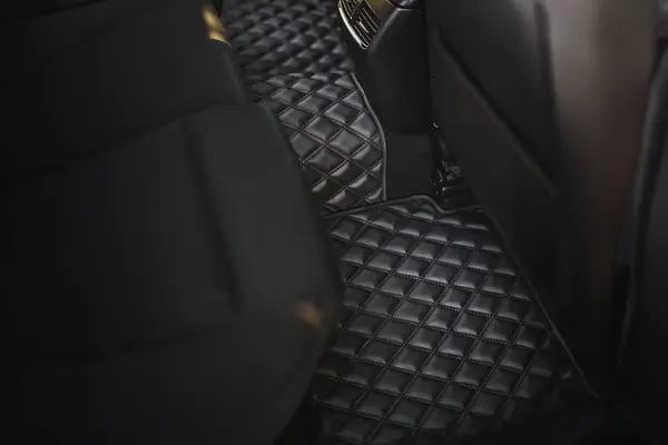 A leather floor mat in the second row of a modern and luxurious car. The interior is black and stylish, with a dashboard that has technology and speed features, a seat, and pedals.