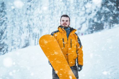 Kış günü snowboard yapmak. Beyaz yamaçta snowboard 'u olan sakallı genç bir adam. Eğleniyor ve adrenalini hissediyor. Arka plan karla kaplı çam ağaçlarının çarpıcı bir görüntüsü..