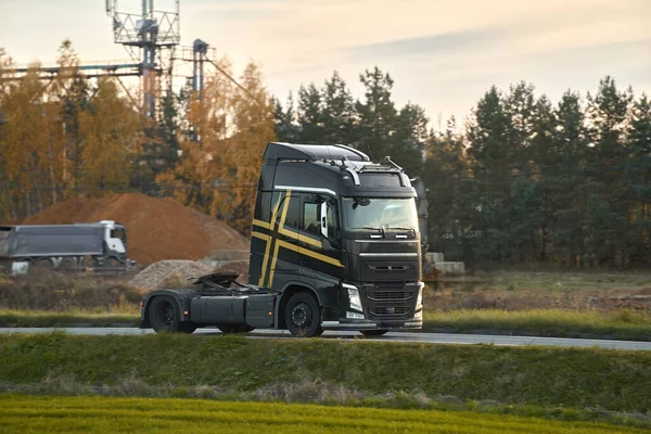 An empty truck with a modern cabin drives on a speedway. The truck has no trailer connected and is carrying no load. The road is bordered by wild grass and a long horizontal structure.
