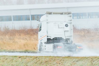 A powerful truck without a trailer speeds along a highway surrounded by industrial buildings and wild grass under a cloudy sky. Sustainable future global shipping.