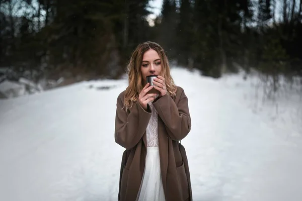 A woman warms her hands with a cup of tea or coffee in the winter nature. She is celebrating and having fun with her partner in the outdoor wonderland.