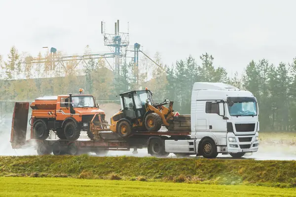 The loader and bulldozer are transported by truck. Heavy construction equipment and trucks on a trailer. Amidst a dusty atmosphere on the highway. White truck transports road working machinery.