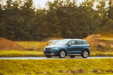 Melez bir SUV arabası görkemli bir doğa ve arka planda altın bir günbatımına sahip manzaralı bir yolda ilerliyor..